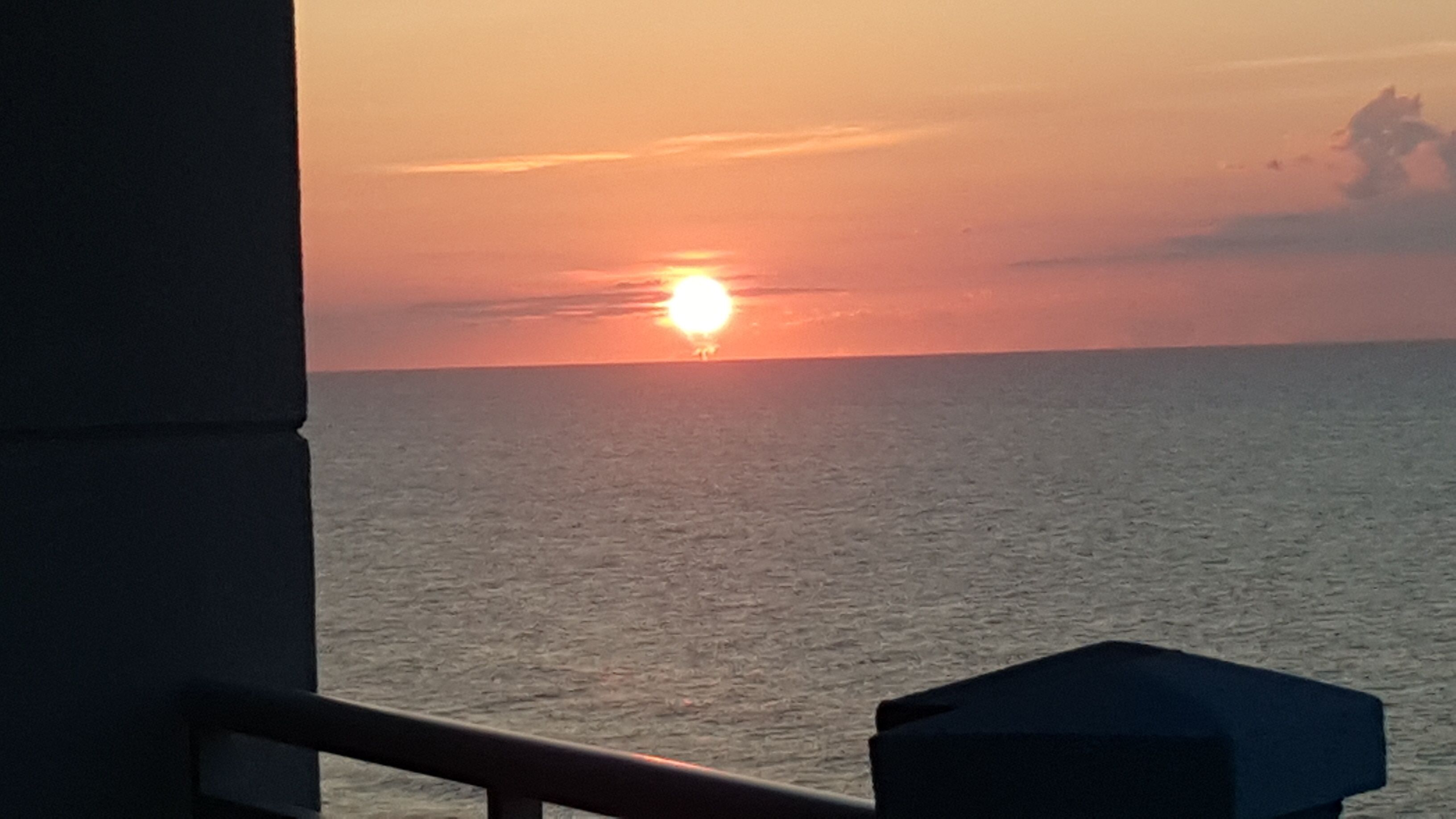 Ein Sonnenuntergang über dem Meer mit einem Balkongeländer und einem Gebäudebau im Vordergrund.