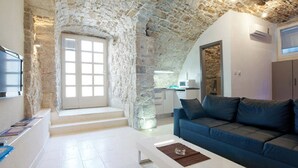 Un salon avec un plafond en pierre, un canapé bleu, un téléviseur à écran plat et une cuisine à l’arrière-plan.