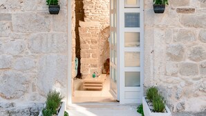 Un bâtiment en pierre avec une porte blanche et des volets verts, un petit marchepied menant à l’entrée, et des plantes en pot de chaque côté.