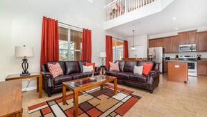 A spacious living room with two leather sofas, a glass coffee table, and a red and beige rug. A kitchen with wooden cabinets and stainless steel appliances is visible in the background. - Serene Pool Home Overlooking Conservation Area 🌳🏊‍♂️ (Davenport)