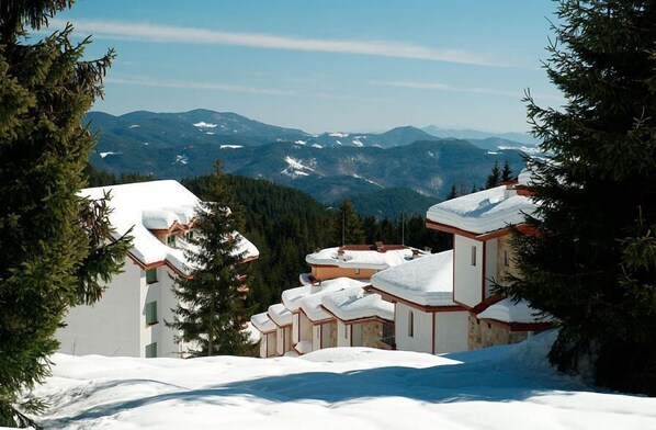 A snow-covered mountain resort with multiple buildings, including a chalet-style structure with a sloped roof, surrounded by evergreen trees.
