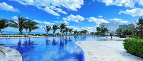 A resort pool area with a clear blue sky, palm trees, and a large building in the background.
