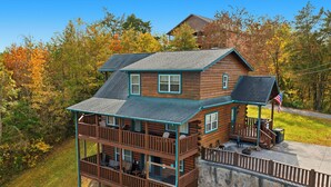 A two-story log cabin with a balcony, a stone retaining wall, and an American flag. - 3BR Cabin Near Dollywood w/ Hot Tub, Game Room & Mountain Views |  Free ticket (Sevierville)