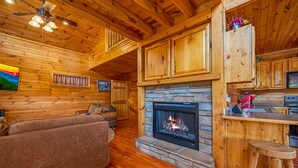 A cozy cabin interior with a stone fireplace, wooden walls, a sofa, and a kitchen area. - 3BR Cabin Near Dollywood w/ Hot Tub, Game Room & Mountain Views |  Free ticket (Sevierville)