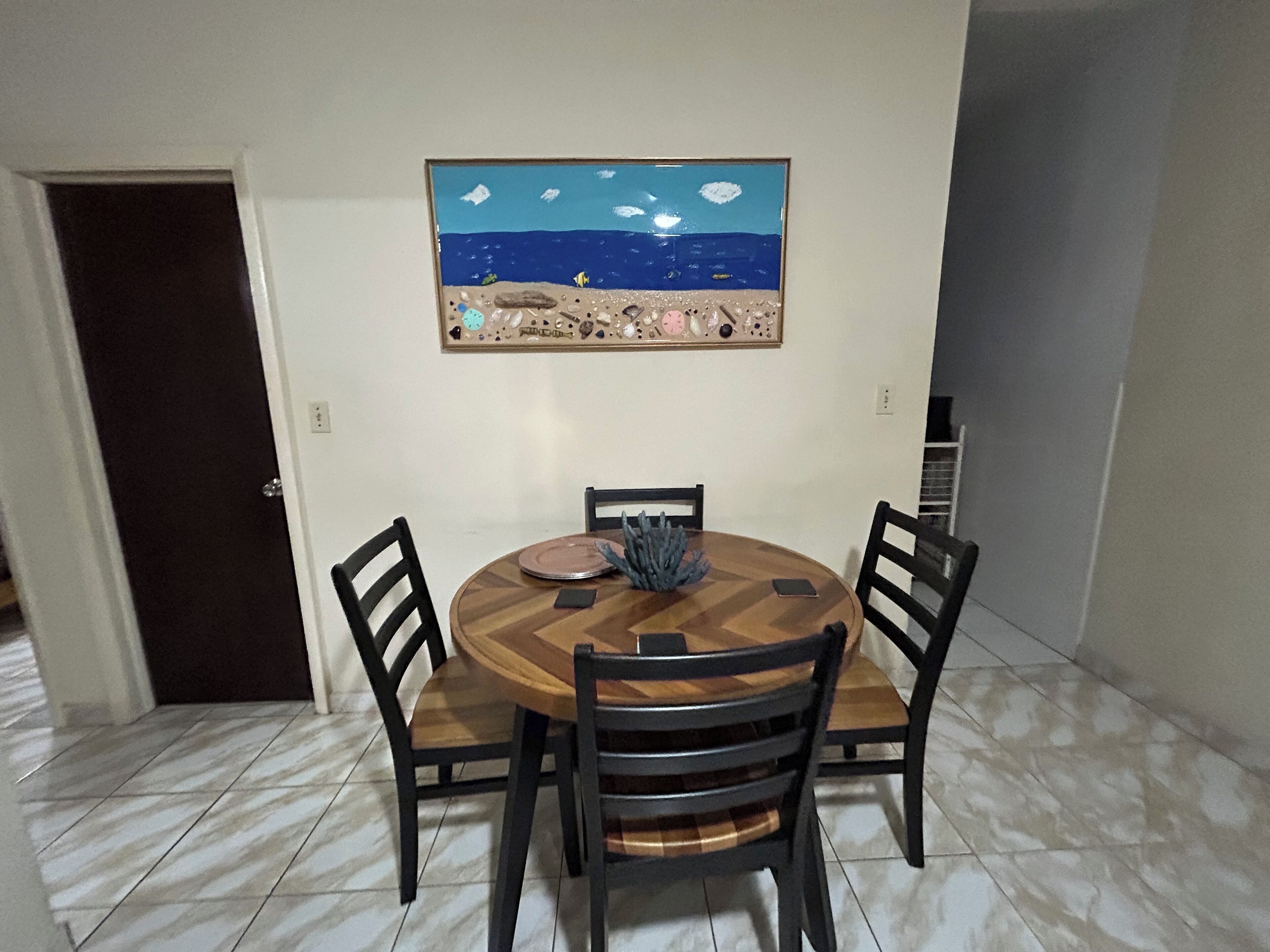 A dining area with a round wooden table, four chairs, and a painting of a beach scene on the wall.