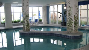An indoor swimming pool with clear blue water, surrounded by white pillars with green plant designs, and a view of the outside through large windows. - Jeffscondos --- Ocean Blue Resort - Ocean Front vacation rental 5bed / 4bath (Myrtle Beach)