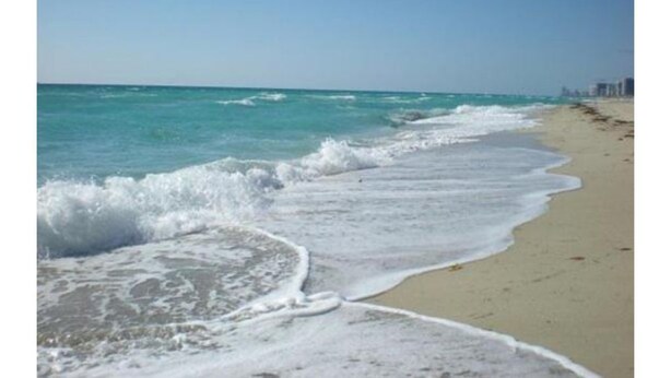 A beach with waves gently lapping onto the shore, a clear sky, and distant buildings. - Jeffscondos --- Ocean Blue Resort - Ocean Front vacation rental 5bed / 4bath (Myrtle Beach)