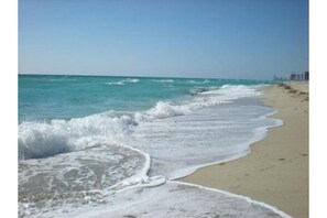 A beach with waves gently lapping onto the shore, a clear sky, and distant buildings.