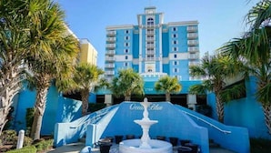 A hotel with a blue facade, a central fountain, and palm trees. - Jeffscondos --- Ocean Blue Resort - Ocean Front vacation rental 5bed / 4bath (Myrtle Beach)