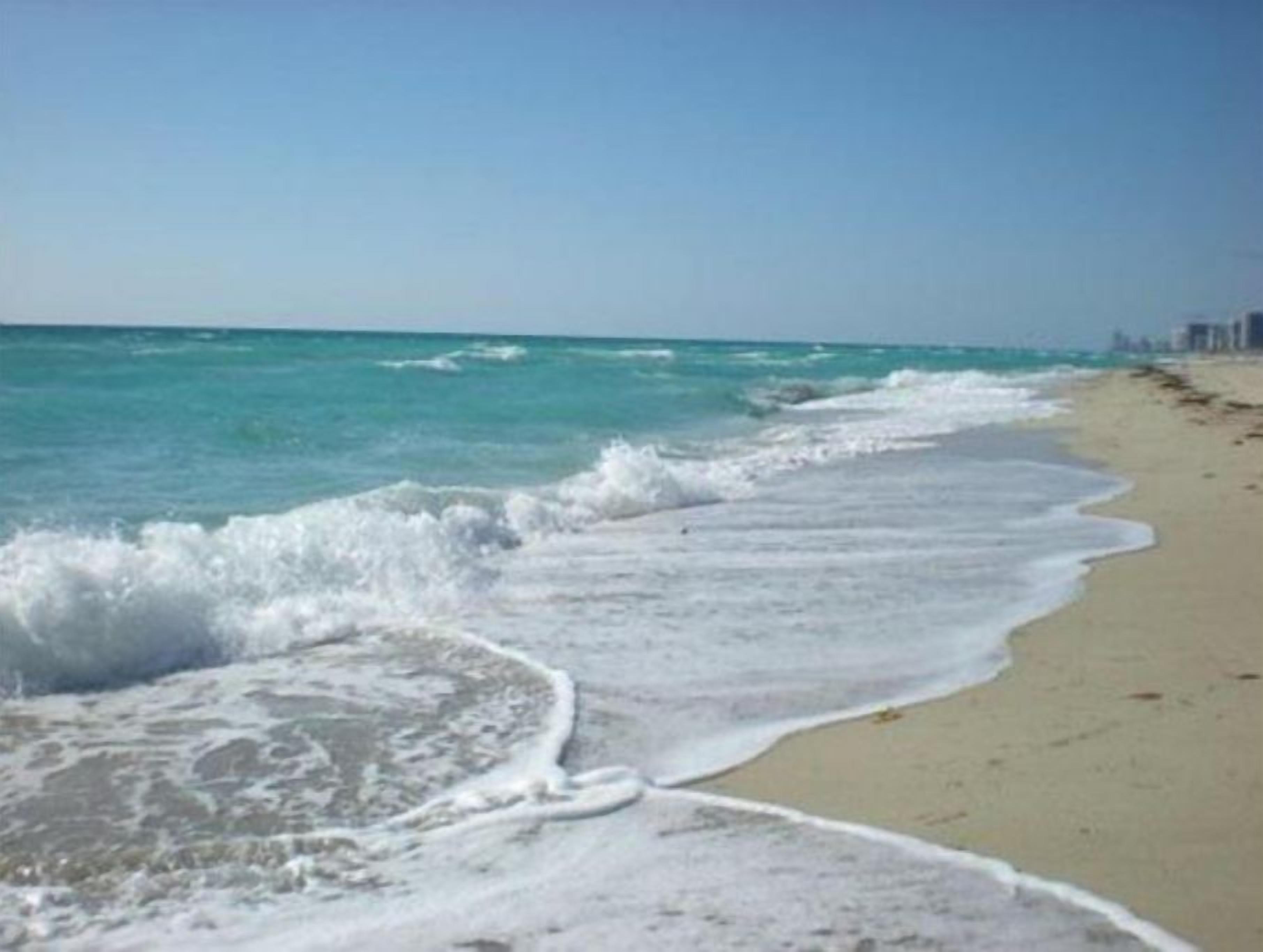 A beach with waves gently lapping onto the shore, a clear sky, and distant buildings.
