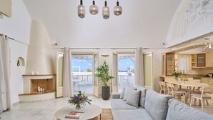 A modern living room with a grey sofa, a coffee table, and a fireplace. There is a dining area with a table and chairs, and a kitchen visible through an open doorway. - La Maison Villa with Large Private Pool - Accommodates 10 (SANTORINI)