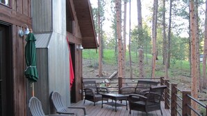 A wooden deck with outdoor furniture, a covered area with chairs, and a view of tall trees in the background.