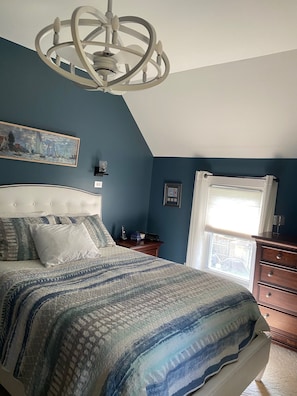 Une chambre à coucher avec un grand lit, un lustre, un tableau au mur, une fenêtre avec des stores et une commode.