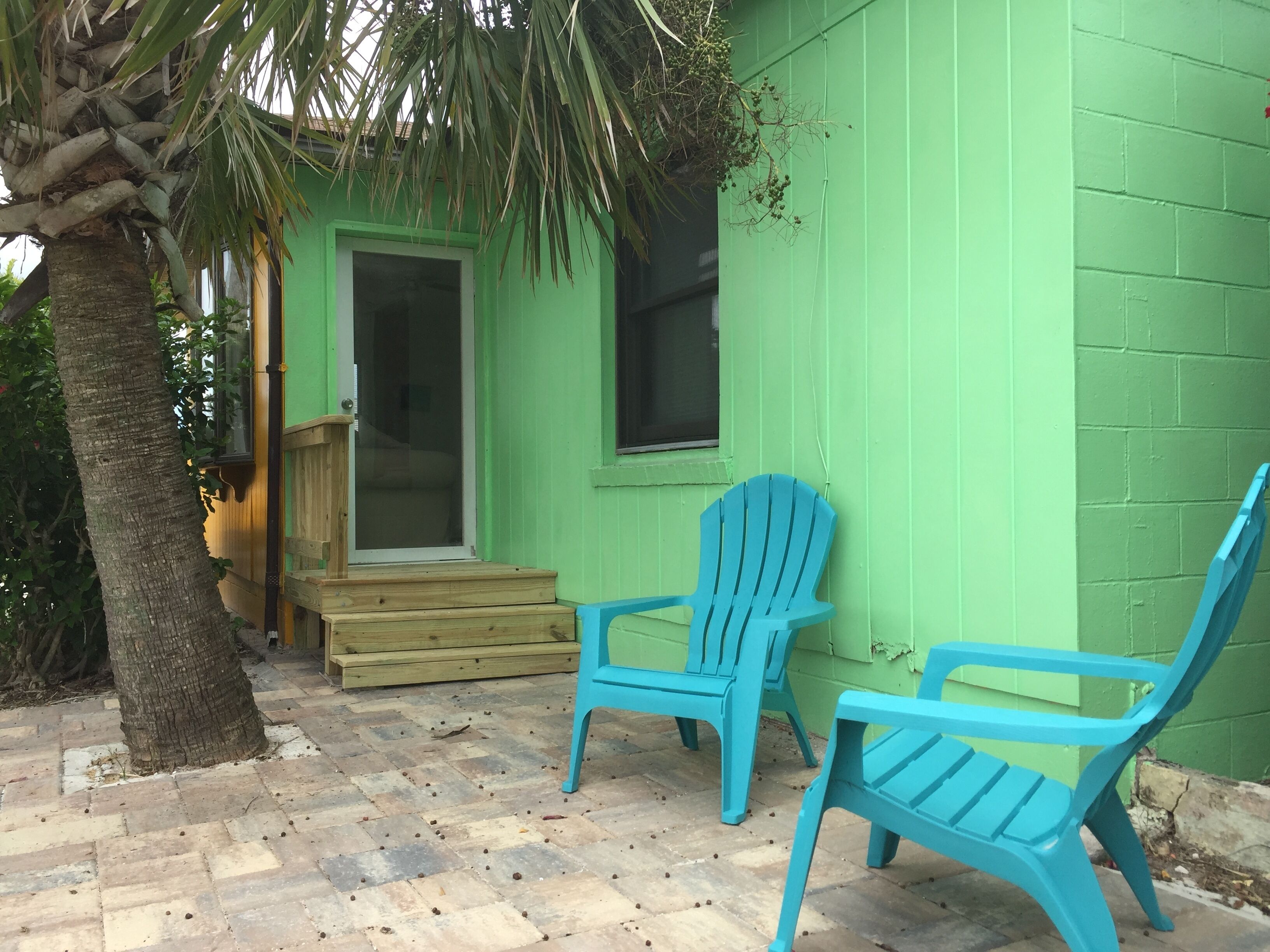 A green building with a wooden porch, two blue chairs, and a palm tree.