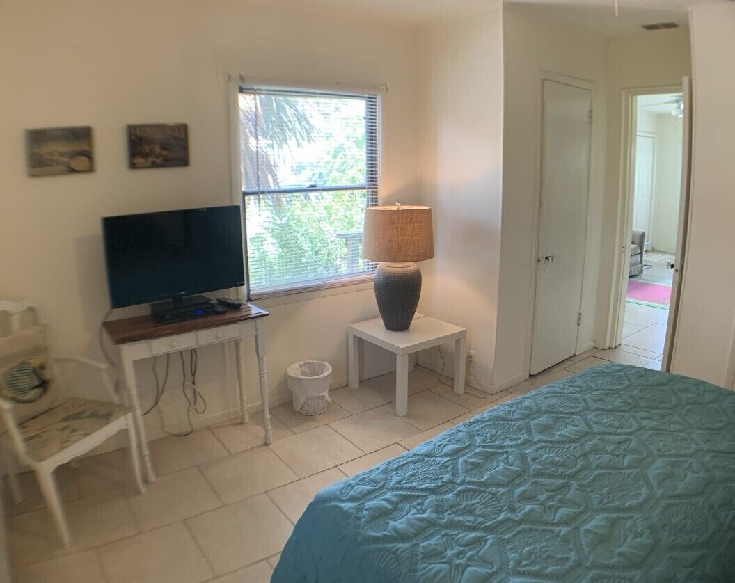 A bedroom with a bed, a desk with a television, a lamp, and a window with blinds.