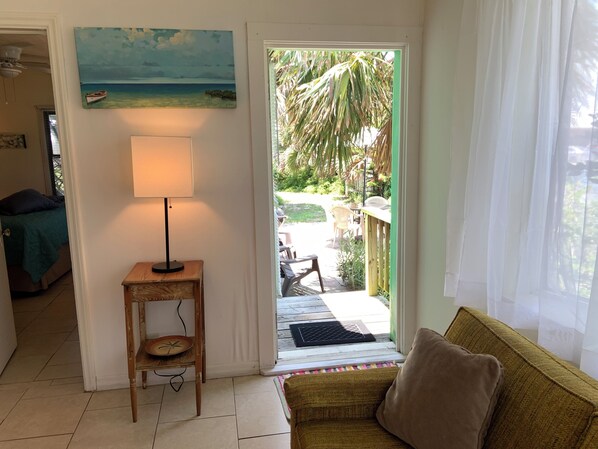 A room with a door leading to an outdoor area, a small table with a lamp, a sofa, and a painting on the wall.