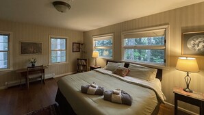 Un dormitorio bien iluminado con una cama grande, dos mesitas de noche, un escritorio y una silla. Hay tres ventanas que ofrecen luz natural y vistas a árboles.