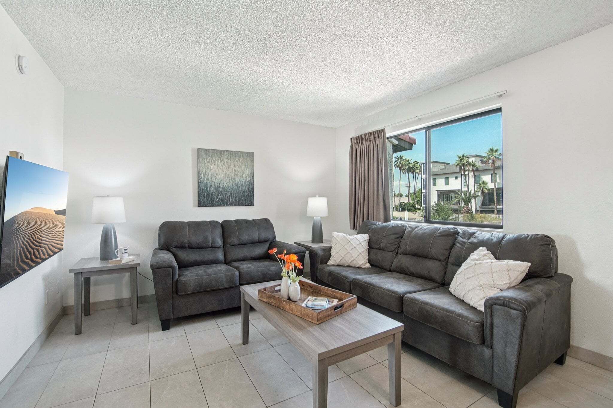 A modern living room with two grey sofas, a coffee table with a tray, and a large window with a view of palm trees.
