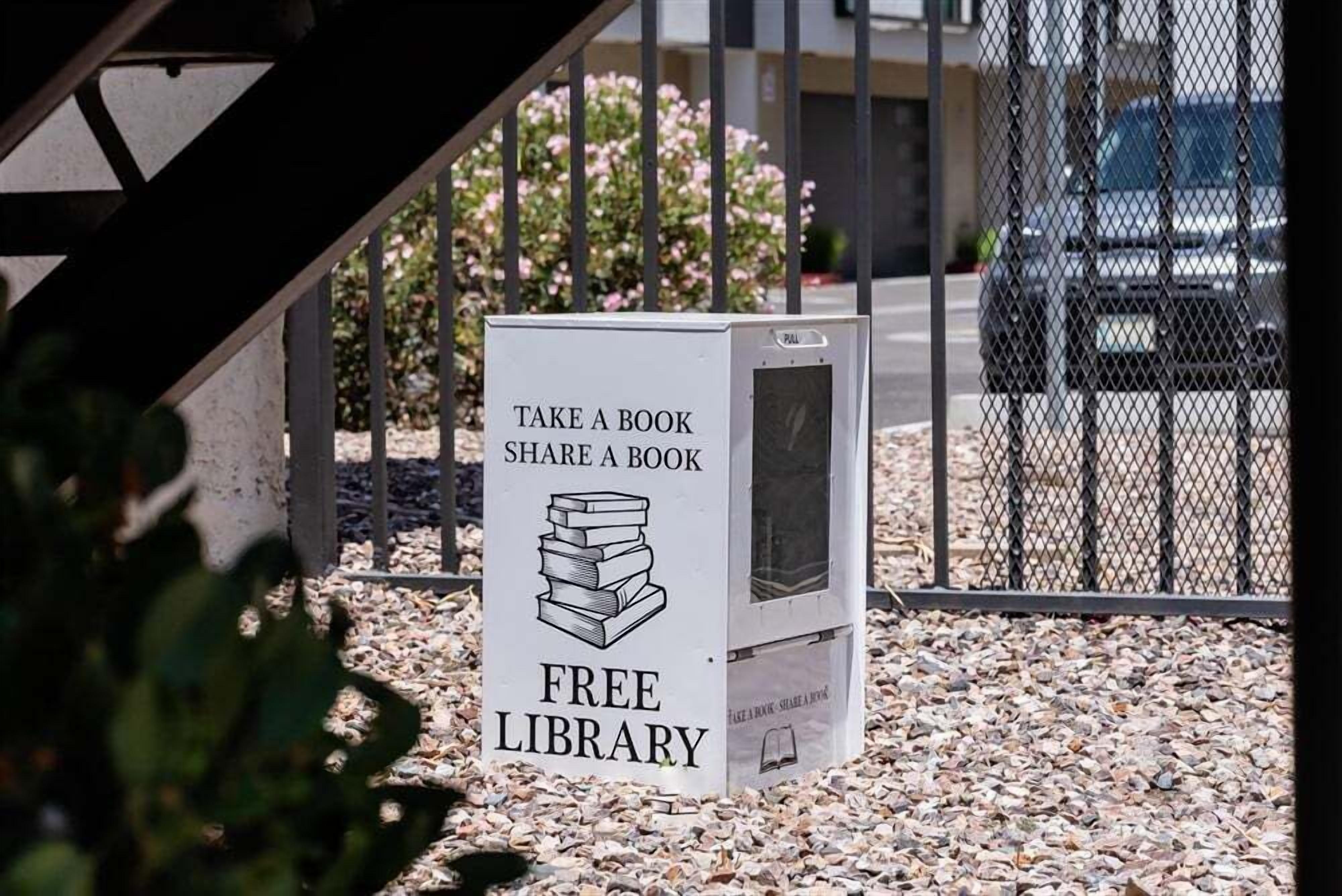 Segnaletica di una biblioteca gratuita con illustrazione di una pila di libri, situata all'aperto su fondo di ghiaia e con una recinzione metallica sullo sfondo.