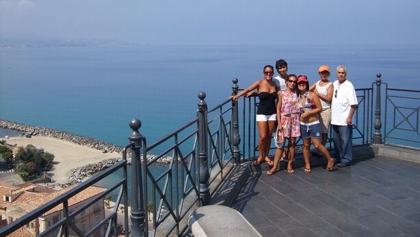 Un grupo de personas de pie en un balcón con vistas a la costa, donde se aprecia un mar despejado y una playa de arena.