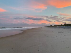 Una playa con un atardecer, una orilla arenosa y algunas personas caminando.