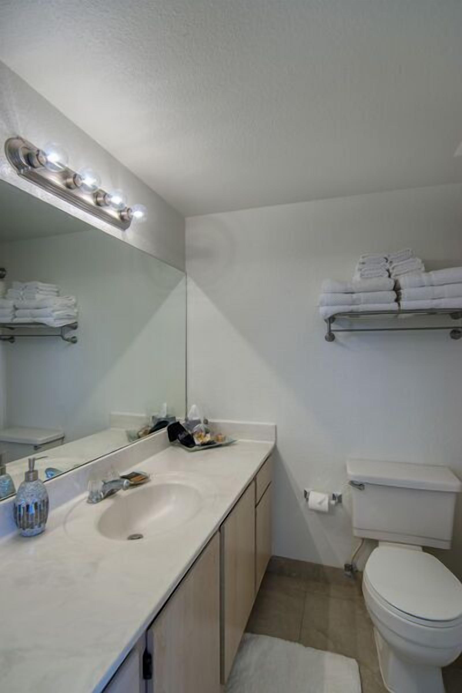A bathroom with a white sink, mirror, and toilet. There are towels on a rack and a shower curtain rod visible.