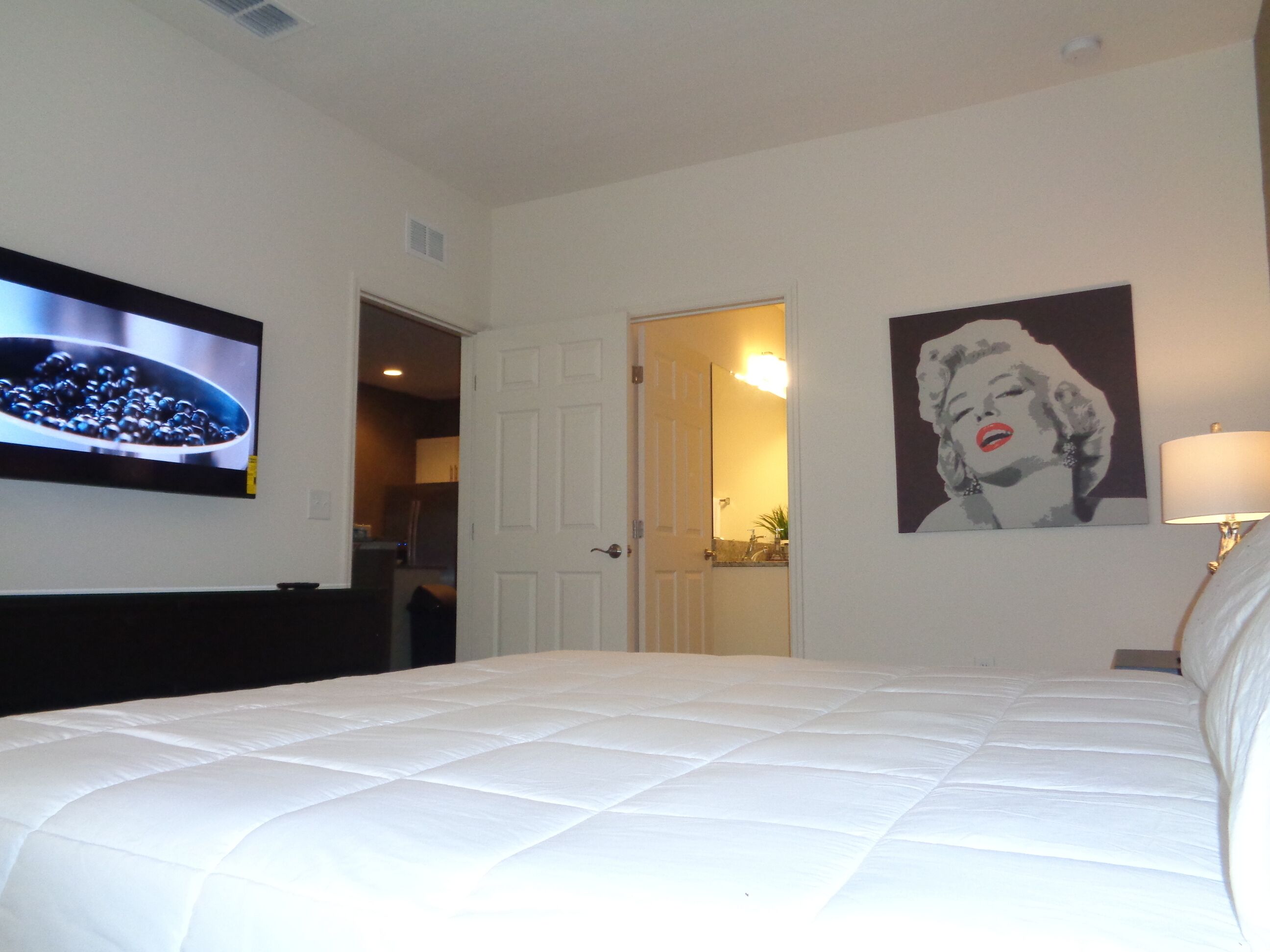 A hotel room with a bed, a television, a door, and a painting on the wall.