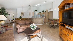 Un salón amplio con un sofá de cuero marrón, una mesa de centro de madera con un centro de mesa floral, un televisor sobre un mueble de madera y una zona de comedor con una mesa y sillas de madera.