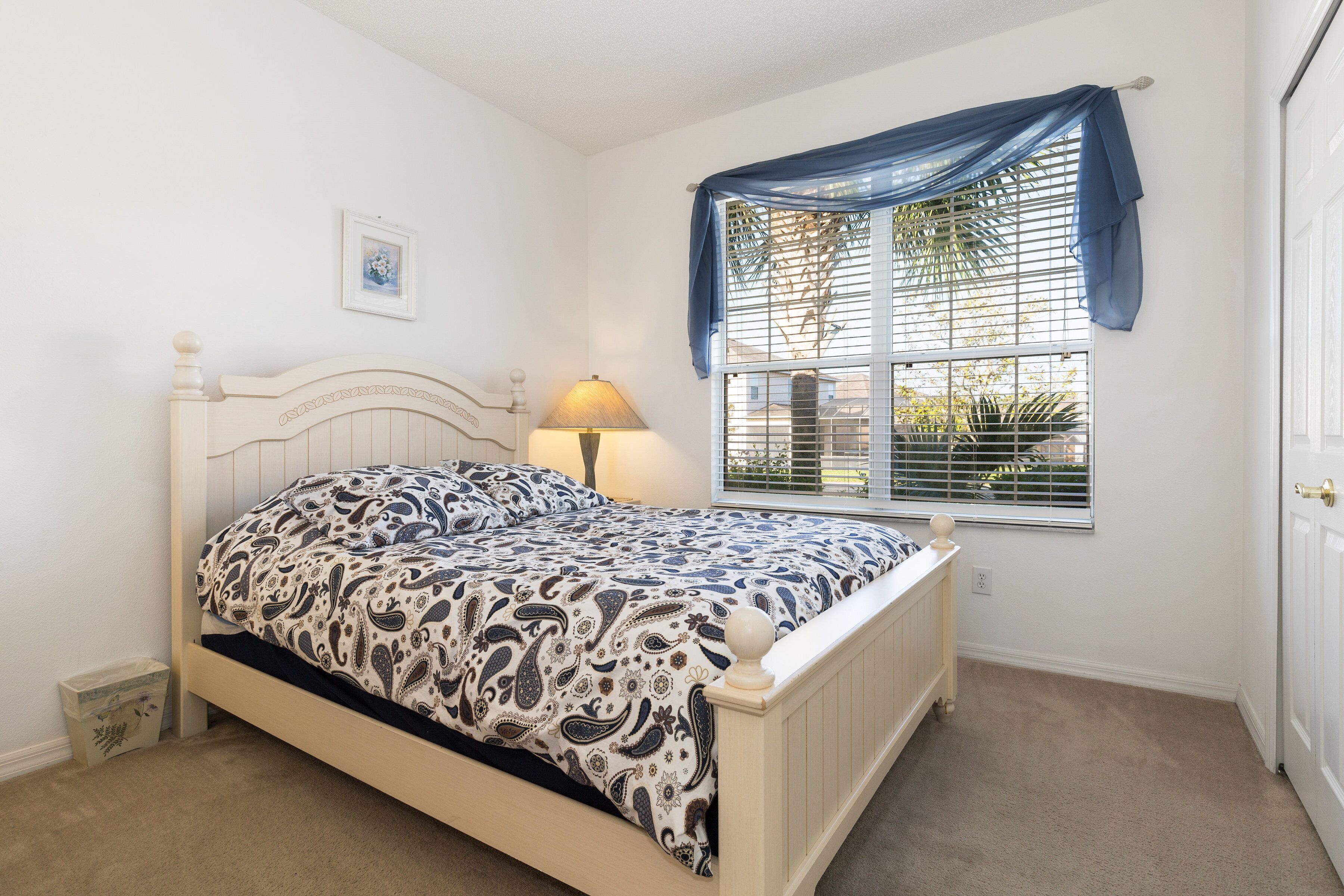 Un dormitorio con cama, ventana con persianas, lámpara y un cuadro en la pared.