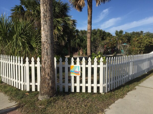 A white picket fence with a colorful artwork attached to it, surrounded by lush greenery and palm trees.