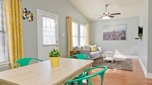 A dining area with a wooden table and green chairs, a gray sofa with cushions, a cityscape painting on the wall, and a ceiling fan. - Spacious 2 Bedroom in Historic Hyde Park (Austin)
