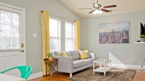 A living room with a gray sofa, yellow curtains, a cityscape painting, a ceiling fan, and a small table with a plant. - Spacious 2 Bedroom in Historic Hyde Park (Austin)