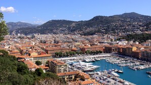 Un port de plaisance où de nombreux bateaux sont amarrés, entouré de bâtiments aux toits de tuiles rouges, serrés les uns contre les autres, le tout se détachant sur un paysage de collines.