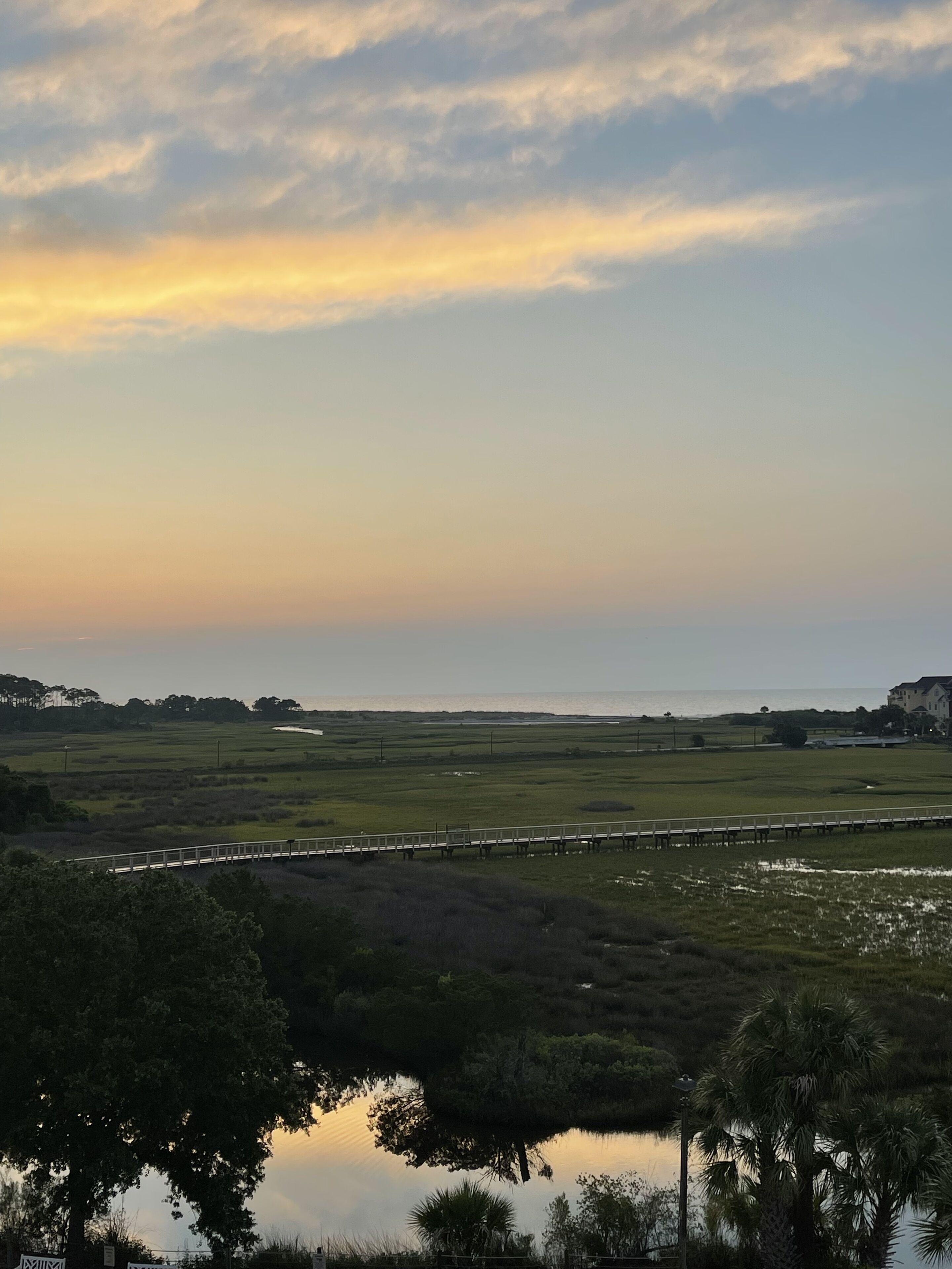 A coastal landscape with a bridge, open fields, and a body of water reflecting the sky.