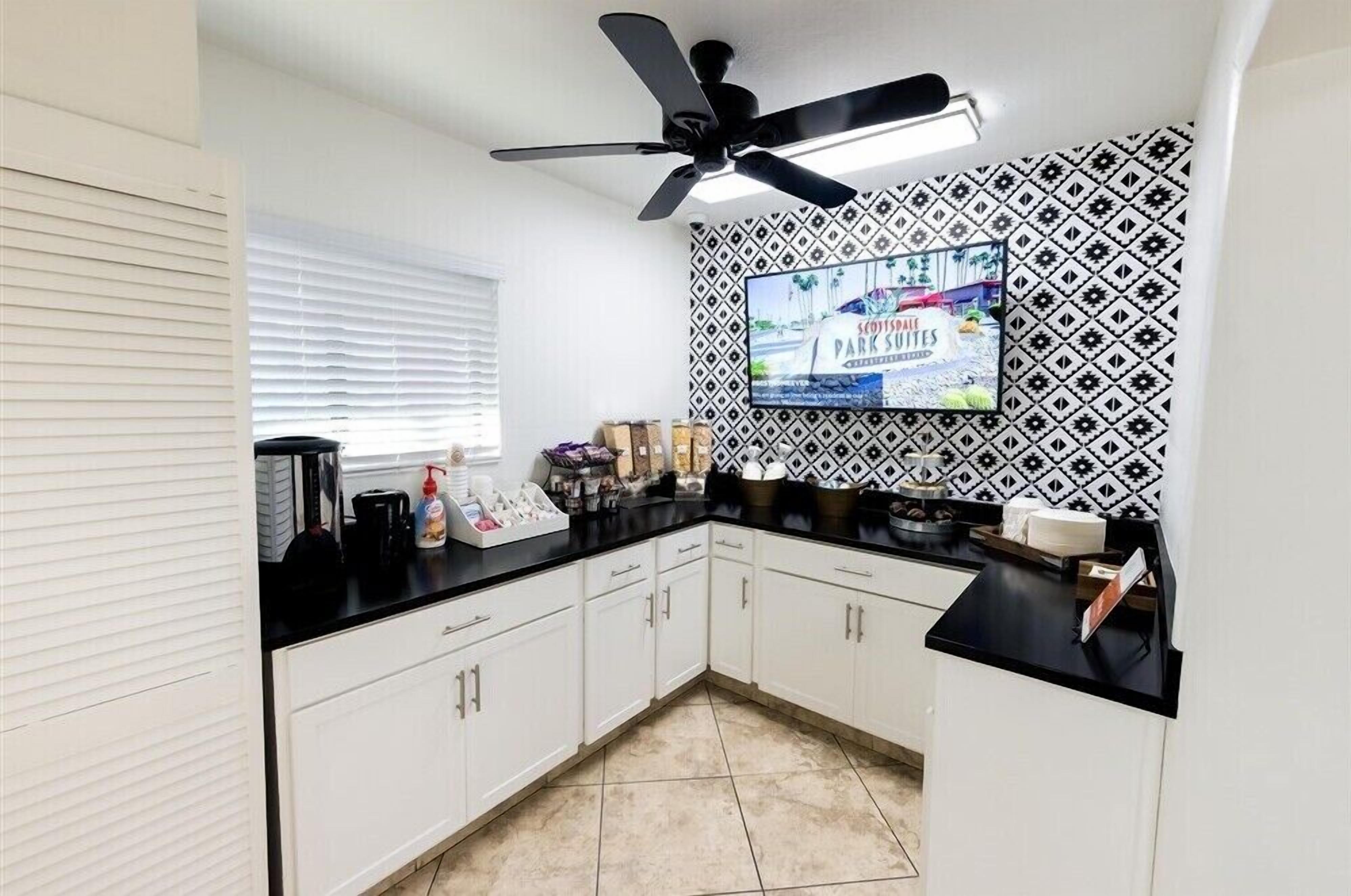 A kitchen with white cabinets, a black countertop, a ceiling fan, and a TV mounted on the wall displaying a scenic view.