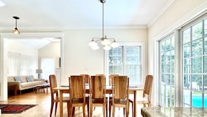 A dining area with wooden chairs and a table, large windows with a view of greenery, and a ceiling light fixture. - Prime East Hampton Village Location. Heated Pool.  Walk To All!  (East Hampton)