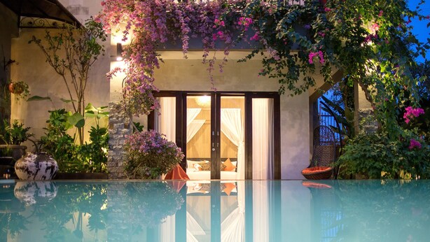 A poolside view of a hotel with a glass-enclosed pool, a stone wall, and lush greenery.