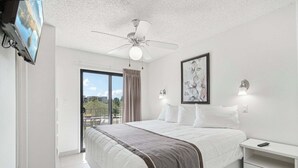 A bedroom with a bed, a ceiling fan, a TV, a painting, and a balcony with a view.