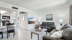 A modern living room with a sofa, a coffee table, and a TV. There is a kitchen area with a microwave and cabinets.