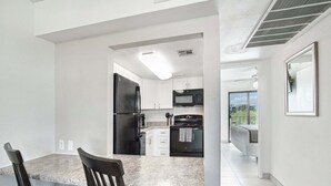 A modern kitchen with white cabinets, a black refrigerator, and a microwave. There is a dining area with a marble countertop and two chairs.