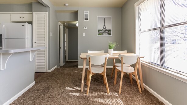 Una cocina compacta con refrigerador, una mesa de comedor con sillas, una planta en maceta y una ventana con persianas.