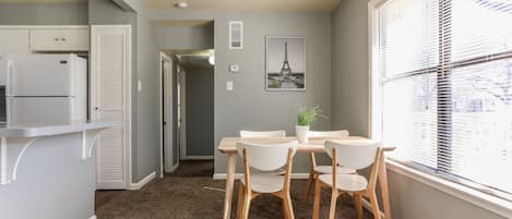 A compact kitchen with a refrigerator, a dining table with chairs, a potted plant, and a window with blinds.