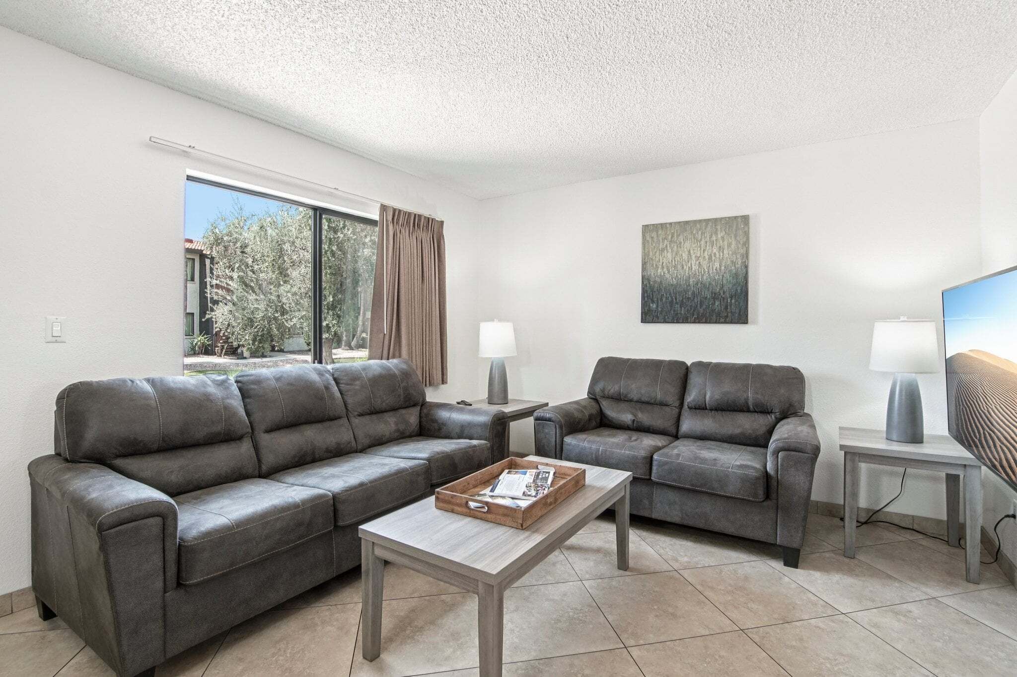A modern living room with two grey sofas, a coffee table, two side tables with lamps, a painting on the wall, and a window with curtains.