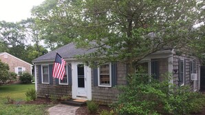 A single-story house with a front porch, a small garden, and an American flag. - Quaint Cape Cod Cottage close to all nantucket sound beaches, and RT28 Fun  (South Yarmouth)