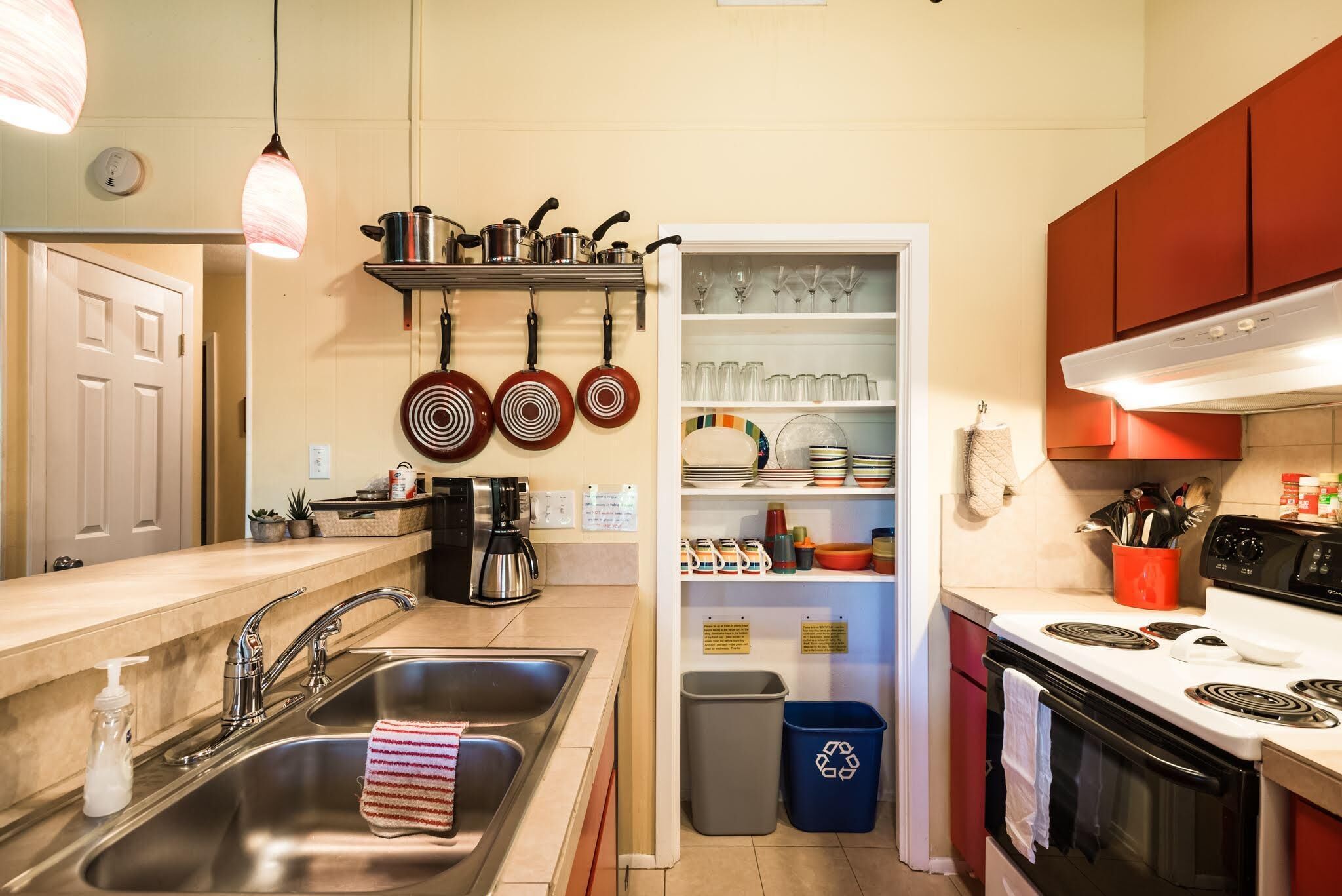 Una cocina con lavamanos de acero inoxidable, cafetera, ollas colgantes y horno empotrado.