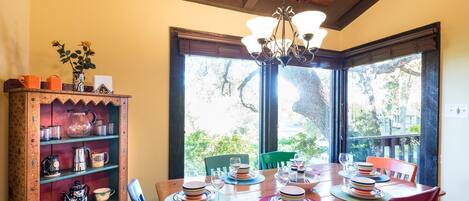 A dining area with a wooden table set for four, surrounded by chairs. A wooden cabinet with shelves is to the left, and a window with a view of greenery is behind the table.