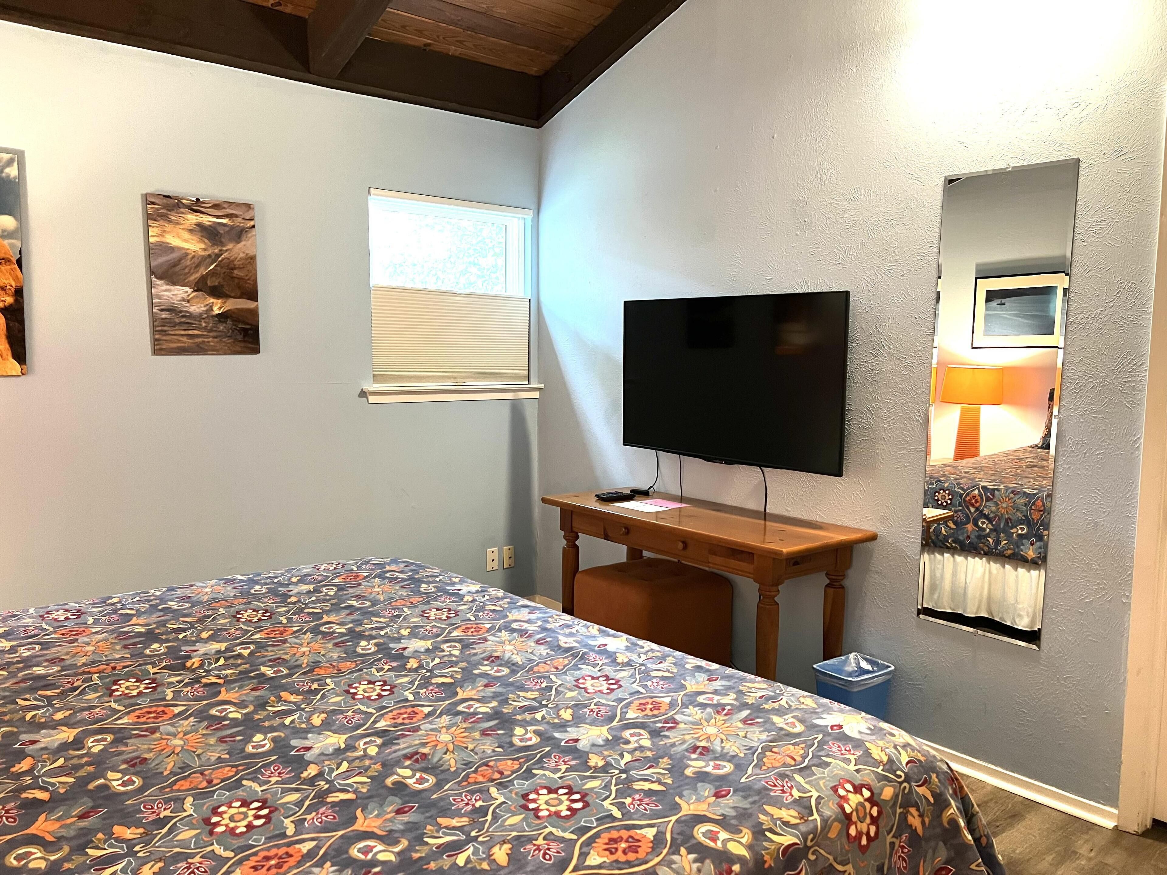 A bedroom with a bed, a desk with a TV, a window with blinds, and a mirror on the wall.