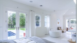 A modern bedroom with a large bed, a freestanding bathtub, and a view of the outdoors through large windows. - Private Westhampton Escape 5BR, Pool, Hot Tub, Near Beach (Westhampton)