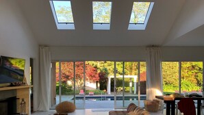 A modern living room with a large sofa, a flat-screen TV, and a view of a garden through large sliding glass doors. - Private Westhampton Escape 5BR, Pool, Hot Tub, Near Beach (Westhampton)