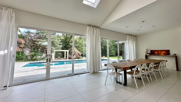 A spacious room with a large dining table, white chairs, and a view of a swimming pool and garden through large sliding glass doors. - Private Westhampton Escape 5BR, Pool, Hot Tub, Near Beach (Westhampton)